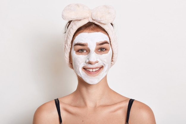 Jeune femme mettant la crème pour le visage tout en posant isolé sur fond blanc, dame robes bandeau avec arc, regardant souriant à la caméra, faisant des procédures de soins de la peau.
