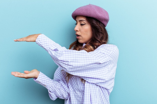 Jeune femme mexicaine isolée sur fond bleu choquée et étonnée tenant un espace de copie entre les mains.