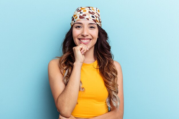 Jeune femme mexicaine isolée sur fond bleu souriante heureuse et confiante, touchant le menton avec la main.