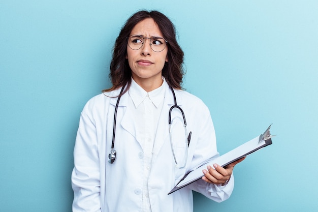 Jeune femme mexicaine médecin isolée sur fond bleu confuse, se sent dubitative et incertaine.