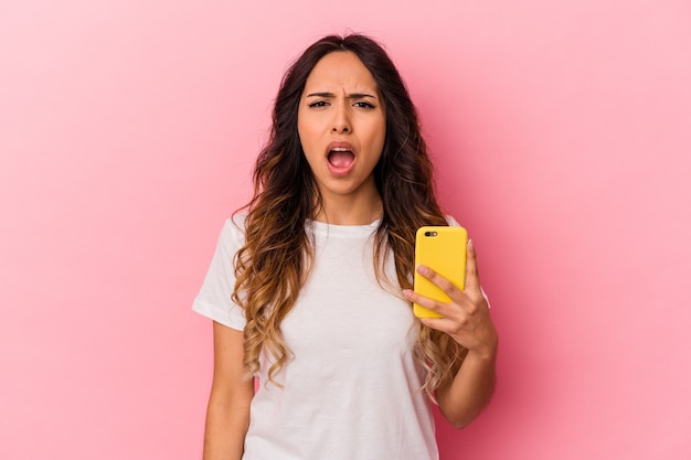 Jeune femme mexicaine tenant un téléphone mobile isolé sur fond rose criant très en colère et agressif.