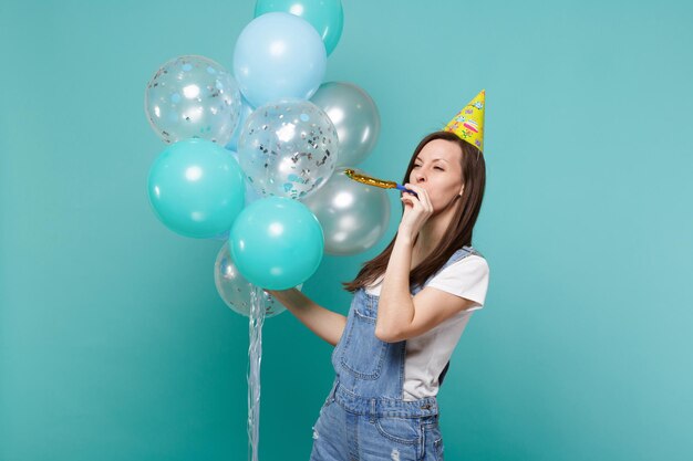 Jeune femme mignonne drôle dans le chapeau d'anniversaire regardant de côté soufflant dans le tuyau, célébrant, tenant des ballons à air colorés isolés sur fond bleu turquoise. Fête d'anniversaire, concept d'émotions de personnes.