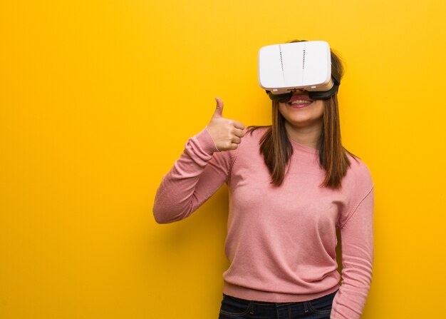 Jeune femme mignonne portant des lunettes de réalité virtuelle souriant et levant le pouce vers le haut