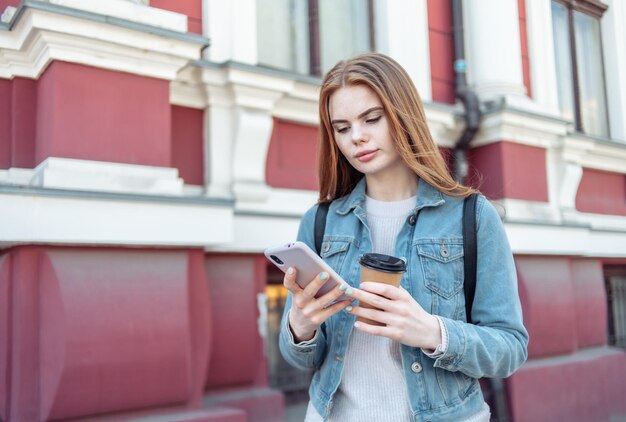 Jeune femme mignonne regarde dans un smartphone et boit du café en déplacement dans la ville