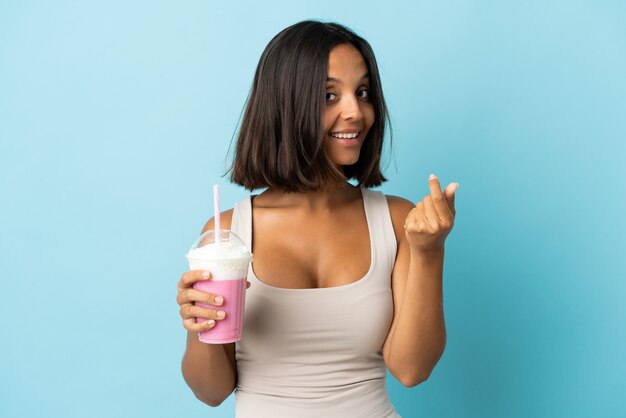 Jeune femme avec milk-shake aux fraises isolé sur mur bleu faisant le geste de l'argent