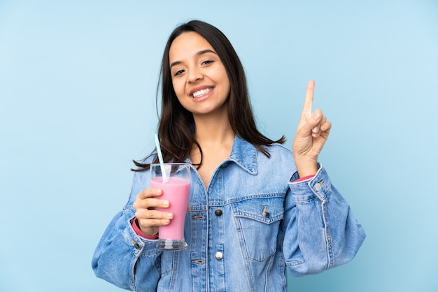 Jeune femme avec milk-shake aux fraises sur mur bleu isolé montrant et levant un doigt en signe de la meilleure