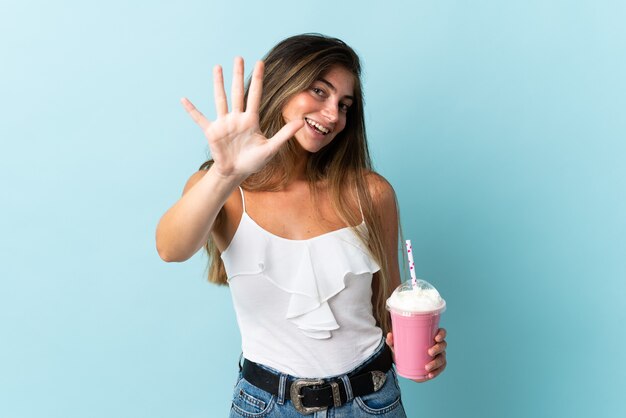 Jeune femme avec milkshake aux fraises isolé sur bleu en comptant cinq avec les doigts