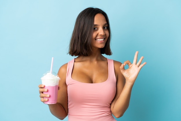 Jeune femme avec milkshake aux fraises isolé sur fond bleu montrant signe ok avec les doigts