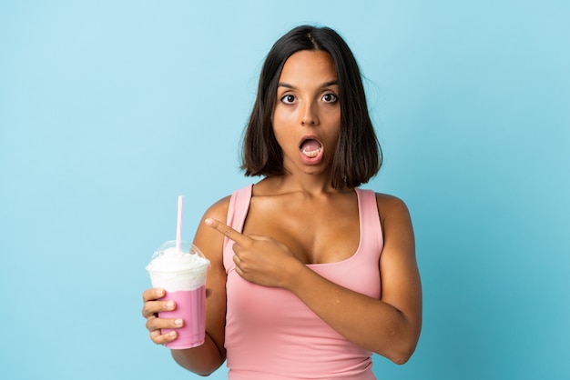 Jeune femme avec milkshake aux fraises isolé sur fond bleu surpris et côté pointant