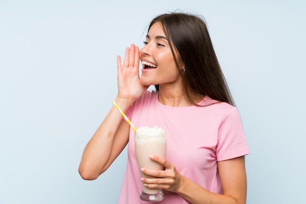 Jeune femme avec un milkshake à la fraise sur fond bleu isolé criant avec la bouche grande ouverte