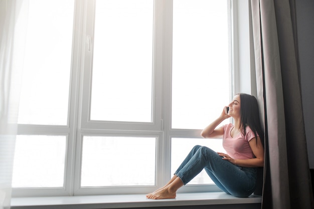 Une jeune femme mince et bien bâtie est assise à la fenêtre et parle au téléphone. Elle lève les yeux. La fenêtre est grande. Elle est assise magnifiquement.