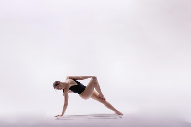 Jeune femme mince en body noir fait des exercices d'étirement de yoga sur fond de mur de studio blanc