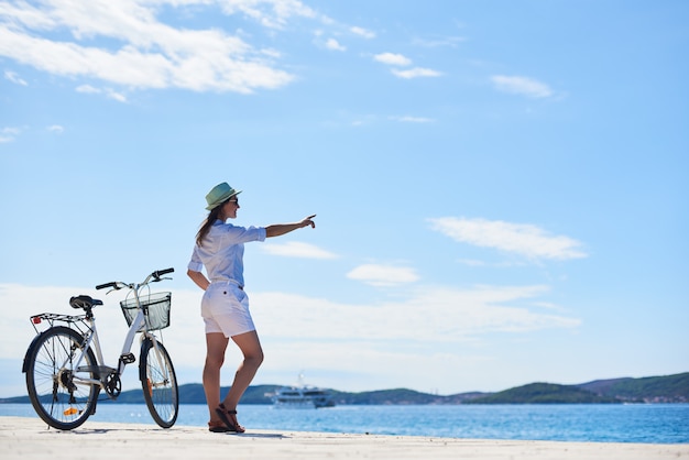 Jeune femme mince debout à vélo sur un trottoir pierreux sous un ciel bleu clair