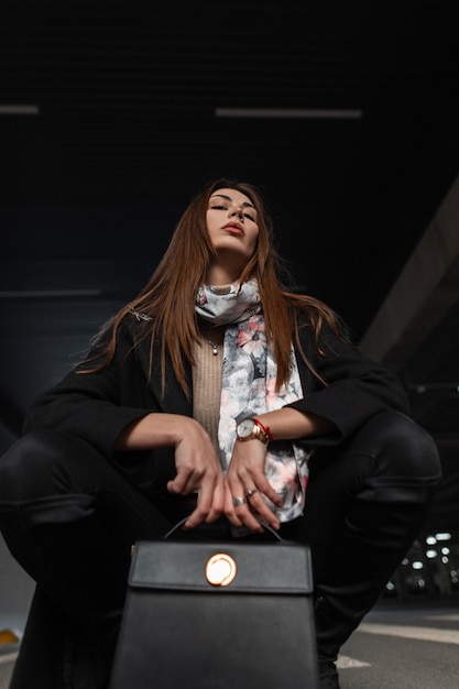 Une jeune femme à la mode aux longs cheveux bruns dans des vêtements de saison noirs élégants avec un sac à main en cuir vintage est assise sur le trottoir de la ville. Jolie fille européenne moderne posant sur la route.