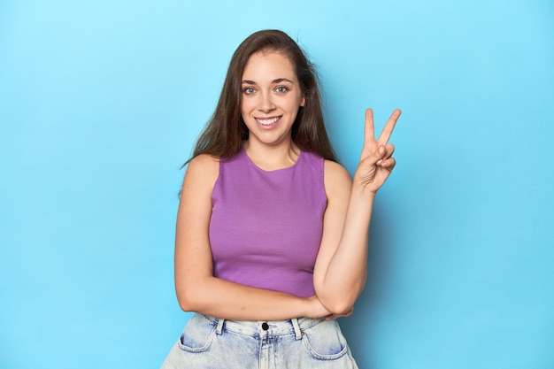 Une jeune femme à la mode dans un haut violet sur fond bleu montrant le numéro deux avec des doigts