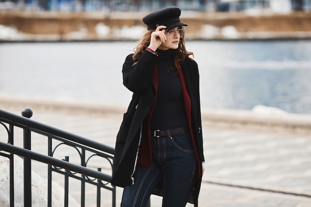 Jeune femme à la mode dans un manteau, un chapeau à la mode et des lunettes de soleil posant sur un fond urbain.