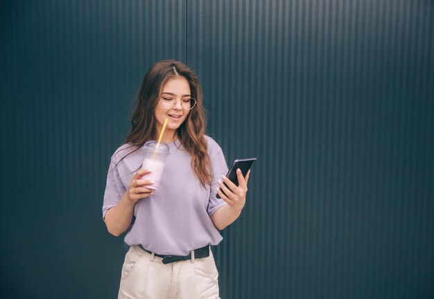 Jeune femme à la mode élégante tenir un cocktail de lait dans la main et regarde le smartphone à travers des verres