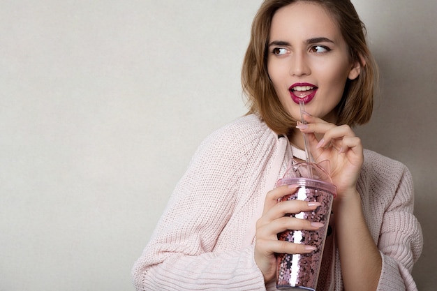 Une jeune femme à la mode porte un pull rose tenant un verre de jus d'orange sur un mur gris. Espace pour le texte