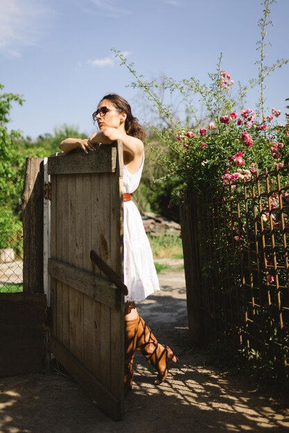 Jeune femme à la mode se tient près de la clôture dans le jardin ou l'arrière-cour de la maison de campagne Heure d'été routine relax concept de récolte