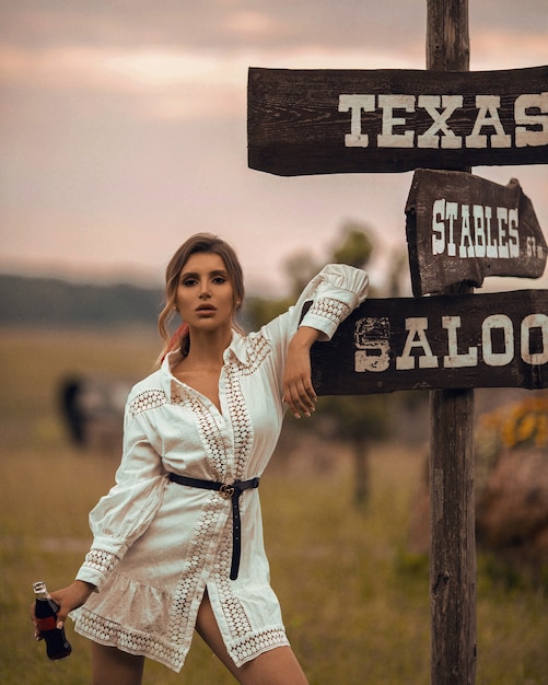 Jeune femme modèle en robe courte blanche posant avec une bouteille de boisson froide au ranch