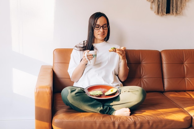 Une jeune femme moderne assise sur le canapé boit du café et mange un sandwich d'avocat sain