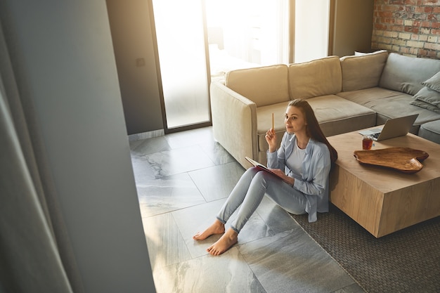 Jeune femme moderne gaie aux pieds nus avec un crayon dans sa main s'asseyant sur un tapis