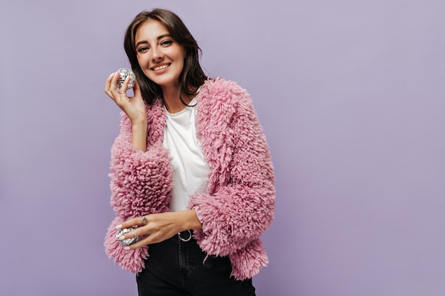 Jeune femme moderne avec un joli sourire en haut blanc pantalon foncé et pull chaud rose regardant dans l'appareil photo et tenant deux petites boules disco