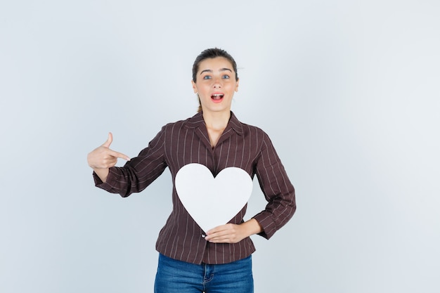 Jeune femme montrant une affiche en papier en chemise, un jean et l'air surpris, vue de face.