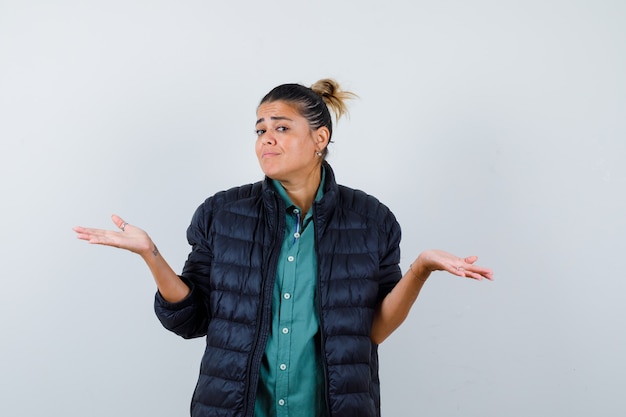 Jeune femme montrant un geste impuissant en chemise, doudoune et semblant confuse. vue de face.