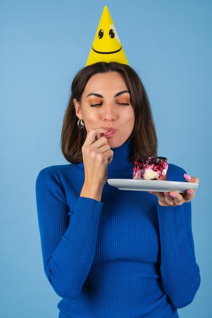 Jeune femme sur un mur bleu fête un anniversaire, tient un morceau de gâteau, heureuse, lèche son doigt avec plaisir