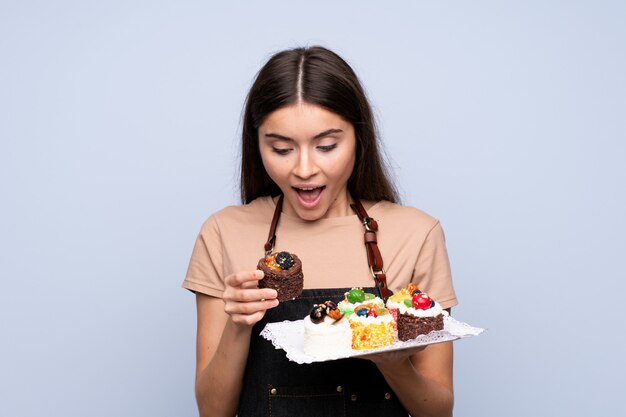 Jeune femme sur mur bleu isolé, tenant des mini gâteaux et surpris