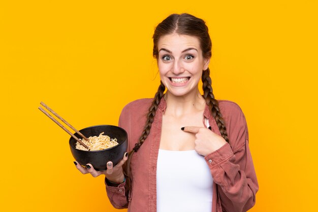 Jeune femme sur un mur jaune isolé avec une expression faciale surprise tout en tenant un bol de nouilles avec des baguettes