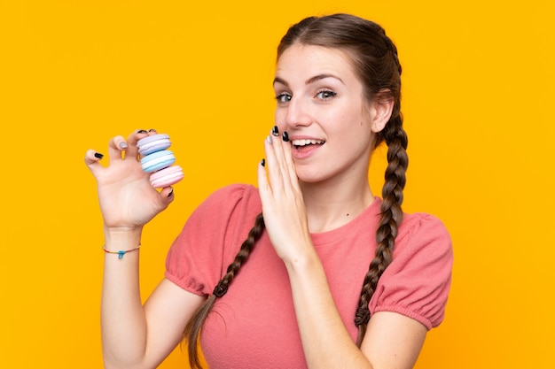 Jeune Femme Sur Un Mur Jaune Isolé Tenant Des Macarons Français Colorés Et Chuchoter Quelque Chose