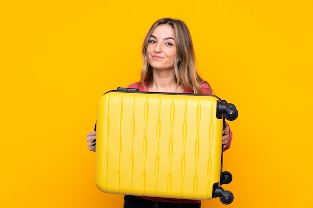 Jeune femme sur un mur jaune isolé en vacances avec valise de voyage