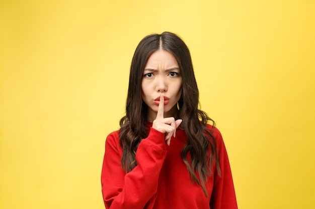 Jeune femme sur un mur jaune montrant un signe de silence geste mettant le doigt dans la bouche.