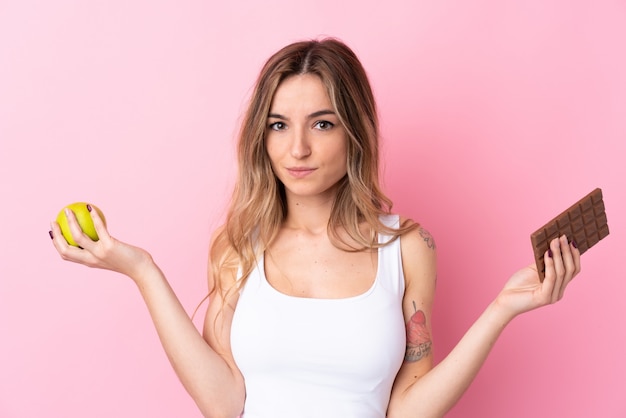 Jeune femme sur un mur rose isolé ayant des doutes tout en prenant une tablette de chocolat dans une main et une pomme dans l'autre