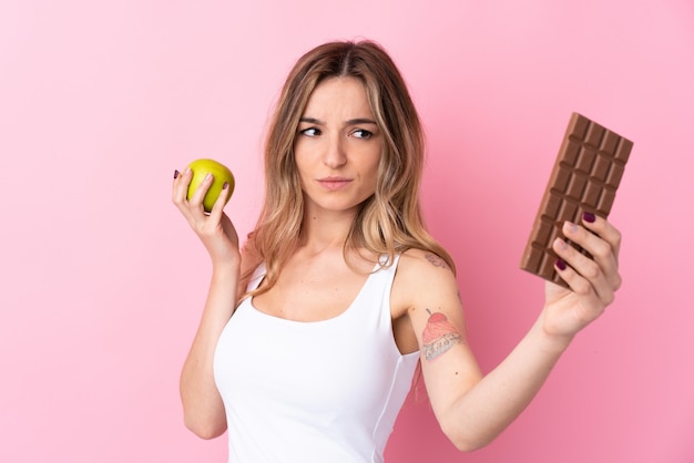 Jeune femme sur un mur rose isolé ayant des doutes tout en prenant une tablette de chocolat dans une main et une pomme dans l'autre