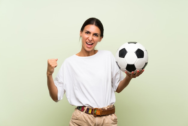 Jeune femme sur un mur vert isolé, tenant un ballon de foot