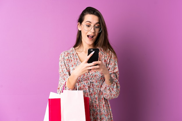 Jeune femme sur un mur violet isolé tenant des sacs à provisions et écrivant un message avec son téléphone portable à un ami