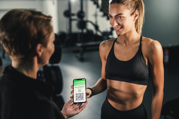 Une jeune femme musclée entre dans une salle de sport et montre à l'entraîneur son passeport de vaccin COVID sur un smartphone.