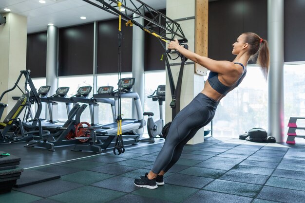 Jeune femme musclée séduisante en vêtements de sport entraîne les muscles avec système de résistance trx dans une salle de sport moderne. Entraînement croisé. Remise en forme moderne