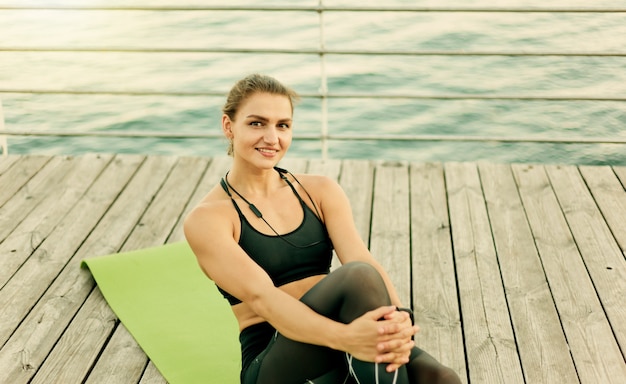Jeune femme musclée en tenue de sport se repose assis sur un tapis à la terrasse de la plage.