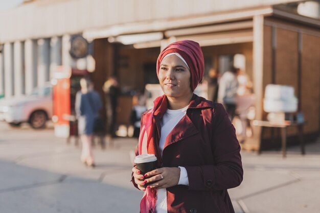 Jeune femme musulmane arabe en hijab marchant au bord de la rue et buvant du café à emporter