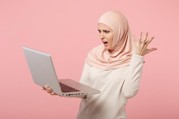 Jeune femme musulmane arabe irritée en vêtements légers hijab posant isolé sur fond rose. Concept de style de vie de l'islam religieux des gens. Maquette de l'espace de copie. Tenir et travailler sur un ordinateur portable.