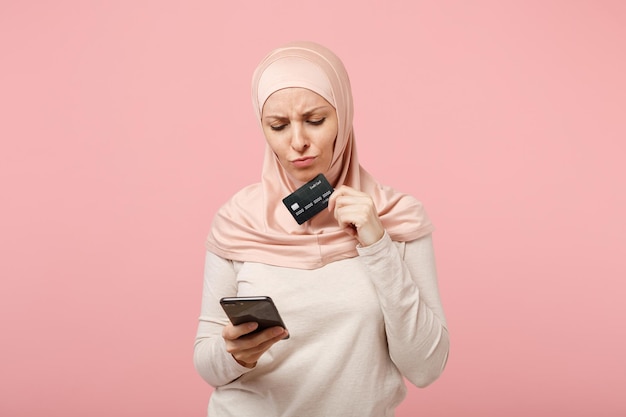 Jeune femme musulmane arabe préoccupée dans des vêtements légers hijab posant isolé sur fond rose. Concept de style de vie de l'islam religieux des gens. Maquette de l'espace de copie. À l'aide d'un téléphone portable, détenez une carte bancaire de crédit.