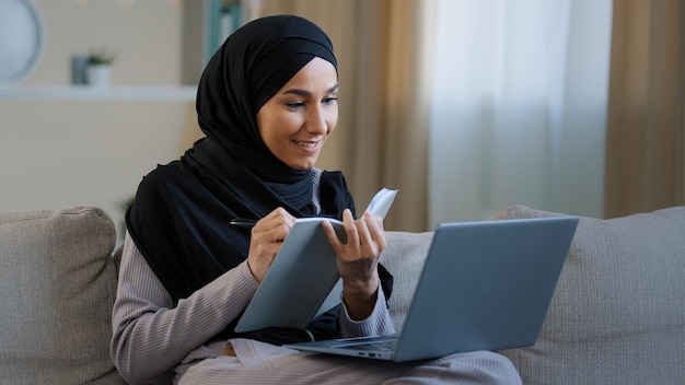 Une jeune femme musulmane arabe s'assoit sur un canapé confortable, une travailleuse indépendante prend des notes dans son journal à l'aide d'un ordinateur portable.