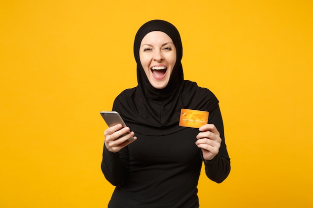 Jeune femme musulmane arabe en vêtements noirs hijab tenir dans la main un téléphone portable, une carte de crédit isolée sur un portrait de mur jaune. Concept de mode de vie religieux des gens.