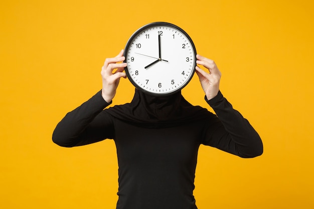 Jeune femme musulmane arabe en vêtements noirs hijab tenir dans les mains une horloge ronde isolée sur un portrait de mur jaune. Mode de vie religieux des gens, concept de gestion du temps.