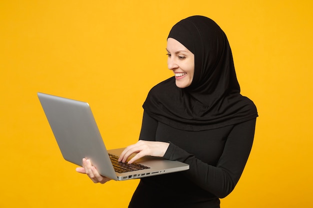 Jeune femme musulmane arabe en vêtements noirs hijab tenir et travailler un ordinateur portable isolé sur un portrait de mur jaune. Concept de mode de vie religieux des gens.