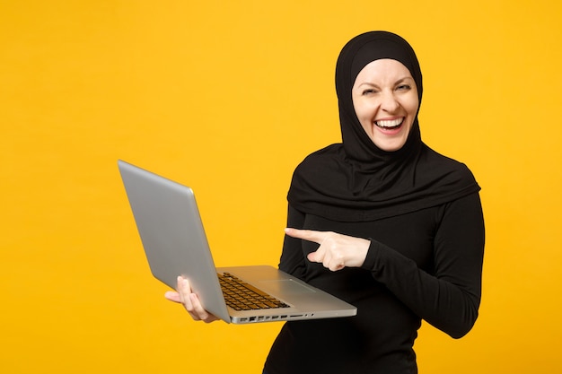 Jeune femme musulmane arabe en vêtements noirs hijab tenir et travailler un ordinateur portable isolé sur un portrait de mur jaune. Concept de mode de vie religieux des gens.